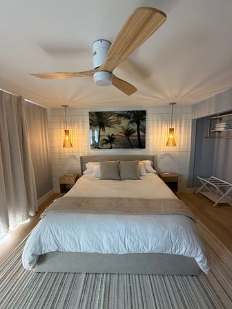 Cozy coastal bedroom with king-size bed, neutral linens, wooden ceiling fan, woven pendant lights, palm beach artwork on shiplap wall, bedside tables and striped rug — bright beachy vibe.