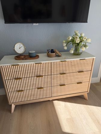 Mid-century modern ribbed wooden dresser under a wall-mounted TV in a sunlit room, topped with a round clock, compact smart speaker on a wooden tray, a woven basket of remotes, and a glass vase of white tulips.