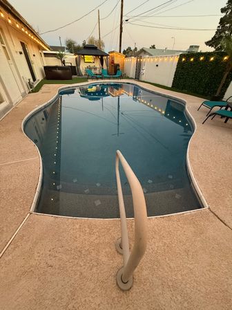 Cozy residential backyard kidney-shaped pool at dusk with handrail and steps in the foreground, mirror-like water reflecting string lights, a small gazebo, teal lounge chairs and patio seating.