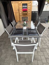 Cozy backyard patio dining area with a glass-top table and grey sling chairs under a curtained gazebo, small potted succulents on the table, a wooden shed with pink flowers and hanging string lights on a paved stone patio.