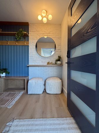 Bright modern entryway with navy beadboard and brass hooks, white painted brick wall topped by a round mirror, twin cream ottomans beneath a slim shelf with potted plants, light wood floors, woven rugs, and a dark wood front door with frosted glass panels.