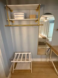 Cozy bedroom dressing nook with gold pipe clothing rack and white shelves holding folded towels and a wicker basket, arched full-length mirror reflecting bed and ceiling fan, white luggage rack on light hardwood floor.