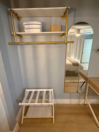 Cozy bedroom dressing nook with gold pipe clothing rack and white shelves holding folded towels and a wicker basket, arched full-length mirror reflecting bed and ceiling fan, white luggage rack on light hardwood floor.