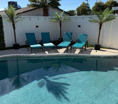 Sunny residential backyard pool with clear blue water, four teal lounge chairs, potted palm trees, white privacy wall and string lights