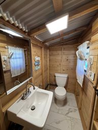 Cozy rustic cabin bathroom with wood-plank walls and corrugated metal ceiling, white sink with chrome faucet and mirror, toilet, marble-look tile floor, LED ceiling light and small curtained window with wall fan.