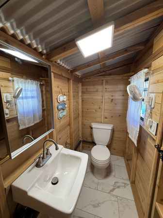 Cozy rustic cabin bathroom with wood-plank walls and corrugated metal ceiling, white sink with chrome faucet and mirror, toilet, marble-look tile floor, LED ceiling light and small curtained window with wall fan.
