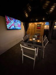 Cozy backyard patio under a covered gazebo with a wall-mounted TV showing basketball, glass-top dining table and chairs, string lights and a wooden shed with potted flowers.