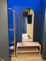 Stylish modern interior entryway with deep navy-blue alcove, arched wall mirror reflecting a hallway door and mounted TV, woven rattan bench and gold metal rolling rack with a woven basket.