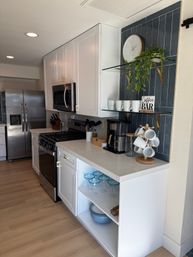 Modern bright home kitchen with white shaker cabinets, stainless steel fridge and gas range, quartz countertop and a cozy coffee bar featuring glass shelves, wooden mug tree, clock and plant against a vertical blue tile backsplash, plus open shelving with blue-rimmed glassware and bowls.
