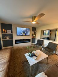 Modern mid-century living room interior with white painted brick media wall, wall-mounted TV above a blue-flame linear electric fireplace, wooden three-blade ceiling fan, marble coffee table with vase, two gray lounge chairs and leather sofa on a patterned rug.
