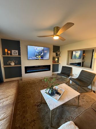 Modern mid-century living room interior with white painted brick media wall, wall-mounted TV above a blue-flame linear electric fireplace, wooden three-blade ceiling fan, marble coffee table with vase, two gray lounge chairs and leather sofa on a patterned rug.