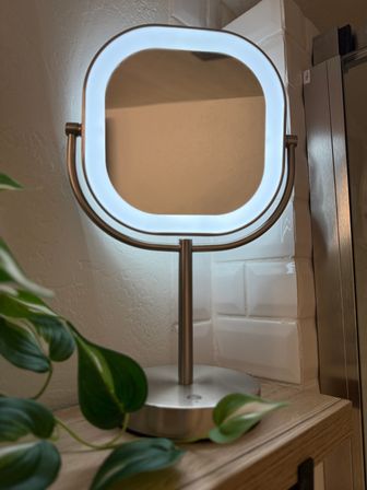 Glowing LED-lit square vanity mirror on a brushed metal stand atop a wooden bathroom counter, framed by white tile and trailing green houseplant