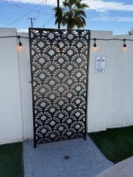 Decorative black metal privacy screen with repeating floral cutouts and an outdoor shower mounted on a white pool wall, warm string lights, pool rules sign, and a palm tree in a sunny backyard.
