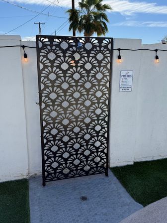 Decorative black metal privacy screen with repeating floral cutouts and an outdoor shower mounted on a white pool wall, warm string lights, pool rules sign, and a palm tree in a sunny backyard.