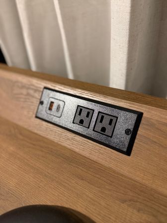 Light wood bedside headboard with a black outlet plate containing two standard electrical sockets and a USB charging port
