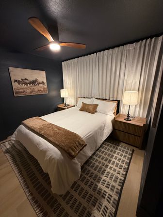 Cozy modern master bedroom interior with dark navy walls, white-dressed bed topped with a brown throw and lumbar pillow, wooden nightstands and lamps, wood-blade ceiling fan, framed horse artwork, textured rug and floor-to-ceiling curtains.