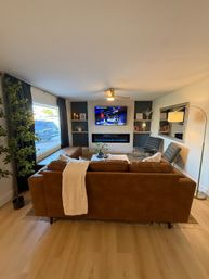 Cozy modern living room with a brown leather sofa and throw facing a wall-mounted TV above a white brick fireplace with a blue linear electric insert, large picture window with dark curtains, gray accent chairs, gold arc floor lamp, indoor plant and light wood floors.