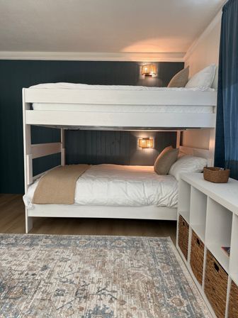 Cozy bedroom with white wooden bunk beds against a navy paneled accent wall, warm woven wall sconces, neutral linens and pillows, wicker storage baskets in a cubby unit and a patterned area rug.