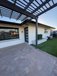 Sunlit modern front patio with dark wood pergola and string lights above gray paver patio, white brick exterior, contemporary frosted-panel gray front door, built-in planter, artificial turf lawn and outdoor seating.
