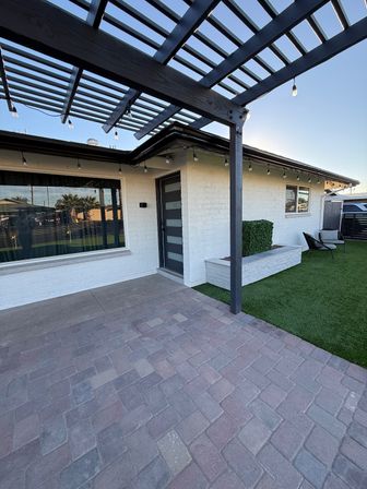 Sunlit modern front patio with dark wood pergola and string lights above gray paver patio, white brick exterior, contemporary frosted-panel gray front door, built-in planter, artificial turf lawn and outdoor seating.