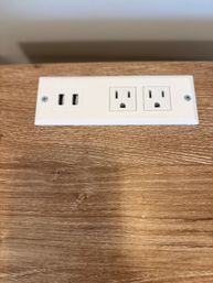White inset power outlet with two USB ports and two standard US sockets mounted on a wood-grain desk surface.