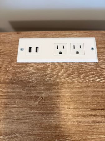 White inset power outlet with two USB ports and two standard US sockets mounted on a wood-grain desk surface.