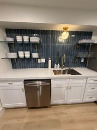 Sleek kitchenette with navy vertical tile backsplash, white shaker cabinets and quartz countertop, brass gooseneck faucet over stainless sink, stainless dishwasher, glass open shelves holding stacked white dishes and glassware, and a warm brass pendant light.