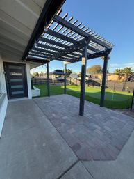 Modern backyard patio with dark wood pergola and string lights over a paver patio, adjacent artificial turf putting green and black fence in a sunny suburban yard with palm trees