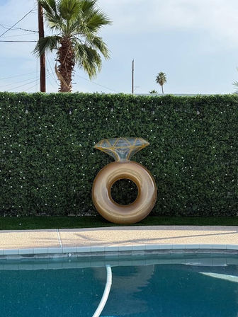 Gold inflatable pool float shaped like a diamond ring leaning against a green hedge beside a backyard pool with palm trees and blue sky