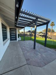 Front patio with dark pergola and hanging string lights over brick pavers, artificial turf putting green and a palm tree in a sunny suburban yard.