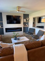 Cozy modern living room with white painted brick fireplace and blue-flame electric insert, wall-mounted TV, brown leather sofa, marble coffee table, gray velvet accent chairs, brass ceiling fan and large mirror.