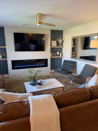 Cozy modern living room with white painted brick fireplace and blue-flame electric insert, wall-mounted TV, brown leather sofa, marble coffee table, gray velvet accent chairs, brass ceiling fan and large mirror.