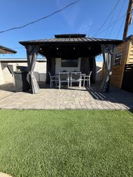 Cozy backyard patio with black metal gazebo and string lights, outdoor dining set on gray pavers, gas grill to the side and green artificial turf under a bright blue sky.