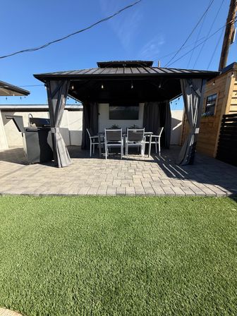 Cozy backyard patio with black metal gazebo and string lights, outdoor dining set on gray pavers, gas grill to the side and green artificial turf under a bright blue sky.