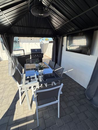 Modern backyard patio under a black metal gazebo with ceiling fan, glass-top dining table with six chairs and succulent centerpieces on stone pavers, wall-mounted TV and stainless gas grill — outdoor dining and entertainment area.