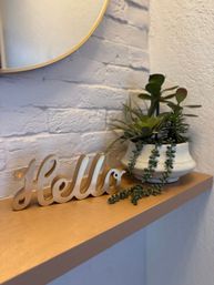 Wooden hello sign on a light wood floating shelf beside a ceramic pot of mixed succulents with trailing string-of-pearls, under a round mirror on a white painted brick entryway wall