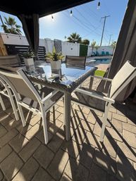Sunny backyard poolside patio with glass-top wicker dining table, mesh chairs, small potted succulents, cabana curtains, string lights and turquoise lounge chairs by the pool