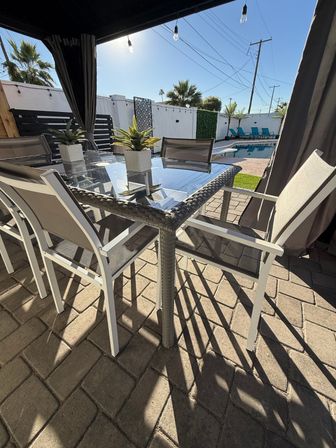 Sunny backyard poolside patio with glass-top wicker dining table, mesh chairs, small potted succulents, cabana curtains, string lights and turquoise lounge chairs by the pool