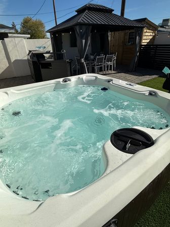 Bubbling backyard hot tub with clear blue water in the foreground, black metal-roof gazebo with outdoor dining set and grill on a sunny patio