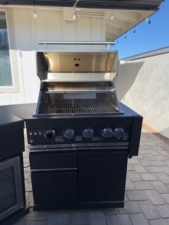 Outdoor stainless-steel gas grill with open hood revealing clean cooking grates, five control knobs and black storage cabinet on a sunlit backyard patio with string lights overhead.