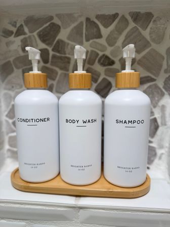 Minimalist bathroom scene: three white refillable dispensers labeled Conditioner, Body Wash, and Shampoo with bamboo pump tops on a wooden tray against a natural stone tile backsplash.