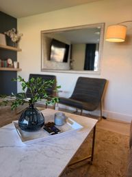 Cozy modern living room: marble coffee table with white tray, dark glass vase of green branches, candle and remotes; two gray lounge chairs, large wall mirror and floor lamp in background.