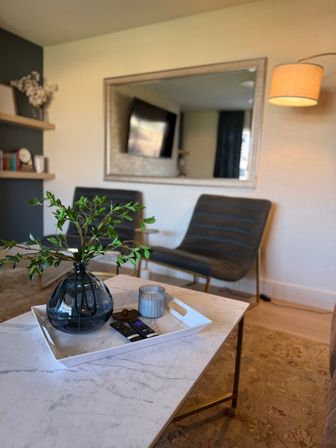 Cozy modern living room: marble coffee table with white tray, dark glass vase of green branches, candle and remotes; two gray lounge chairs, large wall mirror and floor lamp in background.