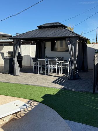 Modern backyard gazebo with black metal roof and curtains, string lights above an outdoor dining table and chairs, grill on a paver patio next to artificial turf under a clear blue sky.