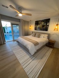 Cozy beach-style bedroom with king bed, neutral linens and throw, striped rug, wood ceiling fan and pendant lights, palm artwork on shiplap wall and sliding glass doors opening to a private pool and lounge chairs.