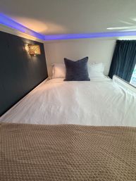 Cozy loft bed top-down view with crisp white bedding, large blue accent pillow, woven beige throw, navy accent wall with rattan wall sconce and soft blue LED cove lighting, dark curtains at the side.