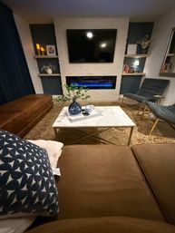 Cozy modern living room with wall-mounted TV above a white-brick electric fireplace with blue flame, marble coffee table with blue vase, brown leather sectional and blue accent chairs.