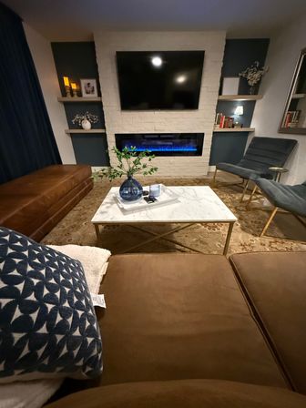 Cozy modern living room with wall-mounted TV above a white-brick electric fireplace with blue flame, marble coffee table with blue vase, brown leather sectional and blue accent chairs.
