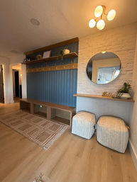 Cozy modern entryway with blue vertical-paneled accent wall, wooden bench cubbies and brass hooks, floating shelves with plants, white brick wall with round mirror and 'Hello' décor, two upholstered ottomans, woven rug and warm wood flooring under a contemporary ceiling light.
