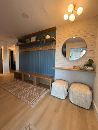 Cozy modern entryway with blue vertical-paneled accent wall, wooden bench cubbies and brass hooks, floating shelves with plants, white brick wall with round mirror and 'Hello' décor, two upholstered ottomans, woven rug and warm wood flooring under a contemporary ceiling light.